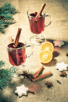 Glass of Christmas mulled wine with cinnamon, star anise and cloves on sackcloth with white biscuits, slices of orange, natural fir tree branches and cones. Color toning effect.