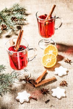 Glass of Christmas mulled wine with cinnamon, star anise and cloves on sackcloth with white biscuits, slices of orange, natural fir tree branches and cones. Color toning effect.