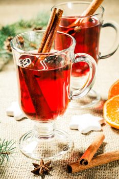 Glass of Christmas mulled wine with cinnamon, star anise and cloves on sackcloth with white biscuits, slices of orange, natural fir tree branches and cones. Color toning effect.