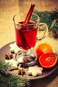 Glass of Christmas mulled wine with cinnamon, star anise and cloves on plate with slices of orange, white biscuits and natural fir tree branches on the background. Color toning effect.