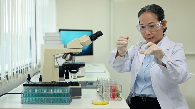 Middle aged female researcher conducting experiment with test tubes and microscope slides in research laboratory.