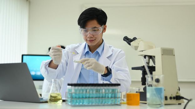 Smiling asian male researcher in white coat conducting experiment with test tubes and microscope slides in research laboratory.