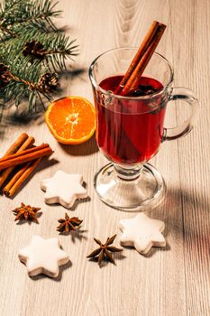 Glass of Christmas mulled wine with cinnamon, star anise and cloves on wooden background with white biscuits, slices of orange, natural fir tree branches and cones. Color toning effect.