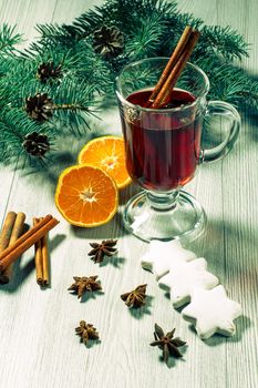 Glass of Christmas mulled wine with cinnamon, star anise and cloves on wooden background with white biscuits, slices of orange, natural fir tree branches and cones. Color toning effect.