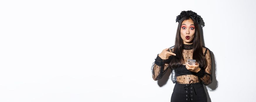 Portrait of amazed asian girl pointing finger at smartphone, showing something about halloween in internet, standing over white background in gothic lace dress.