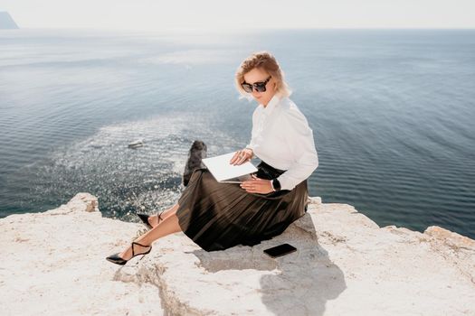 Successful business woman in yellow hat working on laptop by the sea. Pretty lady typing on computer at summer day outdoors. Freelance, travel and holidays concept.