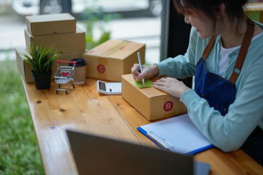 asian entrepreneur working in small start up business online shop company. woman using tape to seal box by packing product to send customers in office. elegant female packaging parcels.