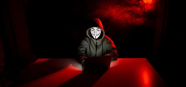 Anonymous in a hood is typing on a laptop in the dark in red smoke