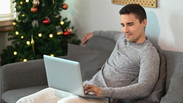 Young man sitting on couch near Christmas tree in living room and using laptop computer.