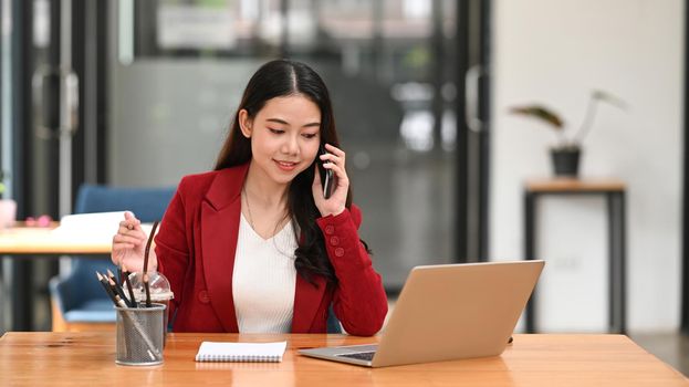 Confident businesswoman sitting at office desk and talking on mobile phone.