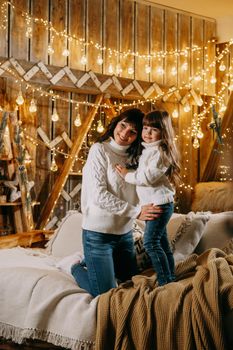 A little girl with her mother in a cozy home environment on the sofa next to the Christmas tree. The theme of New Year holidays and festive interior with garlands and light bulbs