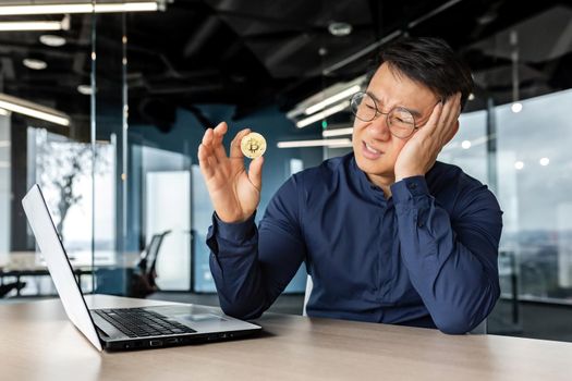 Frustrated asian businessman investor holding crypto currency bitcoin in hands, man working inside modern office building, failure with financial investment.