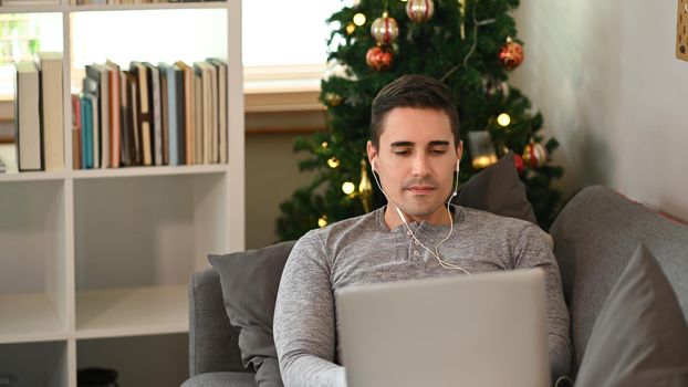 Casual man sitting on comfortable sofa near Christmas tree and using laptop computer.