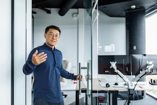 A successful engineer programmer boss invites to his office, an Asian man in a shirt and glasses looks at the camera and smiles and waves to the interlocutor.
