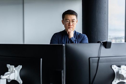 Serious and thinking asian engineer looking at two monitors, close-up photo of office worker, man working inside modern office building, businessman wearing glasses and casual shirt.