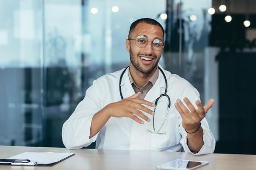 Webcam view, cheerful and successful family doctor consults patients remotely online, video call of man in medical coat smiling and looking at camera, working inside clinic office.