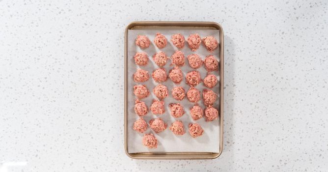 Flat lay. Scooping ground meat with dough scoop into a baking sheet lined with parchment paper to prepare oven-baked meatballs.