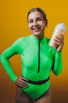 A young smiling blonde in a green swimsuit with a cocktail in her hands is happy about the upcoming vacation, isolated on bright yellow. Summer tropics travel concept.