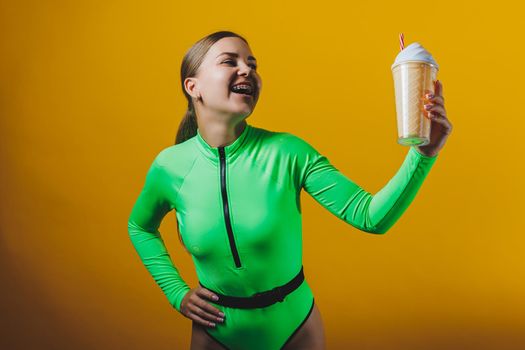 A young smiling blonde in a green swimsuit with a cocktail in her hands is happy about the upcoming vacation, isolated on bright yellow. Summer tropics travel concept.