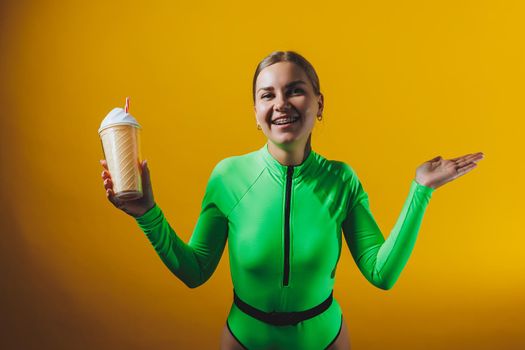 A smiling woman in a green swimsuit with a cocktail in her hands is happy about the upcoming vacation, isolated on a bright yellow background. Summer tropics travel concept.