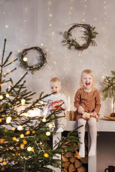 Little brother and sister play on Christmas eve in a beautiful house decorated for the New Year holidays. Children are playing with a Christmas gift. Scandinavian-style interior with live fir trees and a wooden staircase.