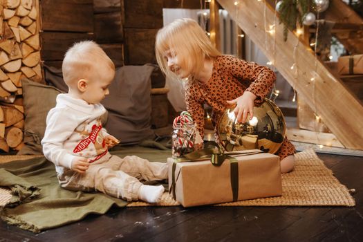 Little brother and sister play on Christmas eve in a beautiful house decorated for the New Year holidays. Children are playing with a Christmas gift. Scandinavian-style interior with live fir trees and a wooden staircase.