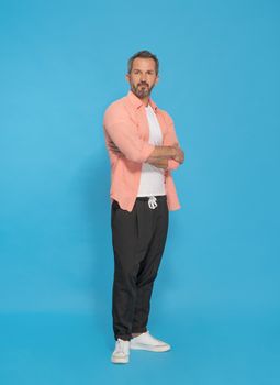 Trendy middle aged grey haired man posing with arms folded looking at camera wearing pink shirt and black jogger pants isolated on yellow background. Mature fit man, health lifestyle concept.