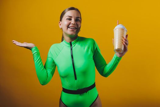 A young smiling blonde in a green swimsuit with a cocktail in her hands is happy about the upcoming vacation, isolated on bright yellow. Summer tropics travel concept.