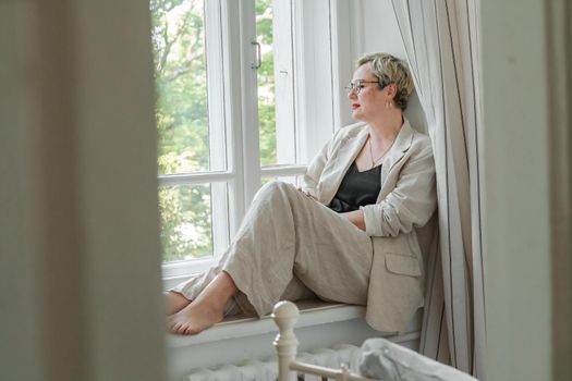 A middle-aged woman in a beige suit and black tank top sits mysteriously and looks out the window on the windowsill. Green trees outside.