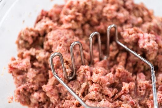 Mixing ingredients with a potatoe masher in a glass mixing bowl to prepare oven-baked meatballs.