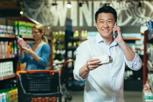 A young handsome Asian man stands at the department of household chemicals, chooses pads in the supermarket . He calls his wife, girlfriend, consults, asks, smiles.