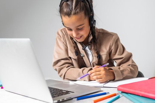 Smiling little Caucasian girl in headphones handwrite study online using laptop at home, cute happy small child in earphones take Internet web lesson or class on computer, homeschooling concept.