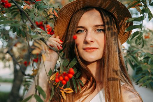 A beautiful blonde girl in a hat with long hair, on a walk in the autumn season. Portrait of a woman at a rowan tree.