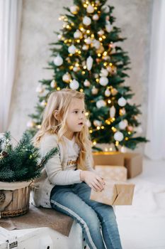 A blonde girl with a Christmas gift in her hands, on the background of a Christmas tree.The concept of New Year's decor and New Year's holidays.