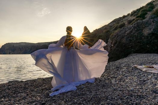 A mysterious female silhouette with long braids stands on the sea beach with mountain views, Sunset rays shine on a woman. Throws up a long white dress, a divine sunset