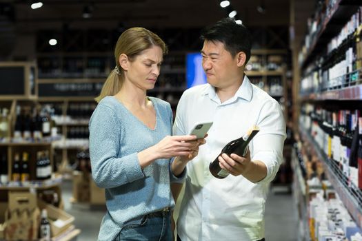 Dissatisfied international couple, Asian man and woman buying wine. They are standing in the alcohol department in the supermarket, unable to choose a wine, holding a bottle and a phone, arguing.