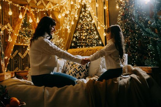 A little girl with her mother in a cozy home environment on the sofa next to the Christmas tree. The theme of New Year holidays and festive interior with garlands and light bulbs.