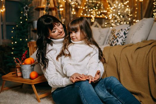 A little girl with her mother in a cozy home environment on the sofa next to the Christmas tree. The theme of New Year holidays and festive interior with garlands and light bulbs.