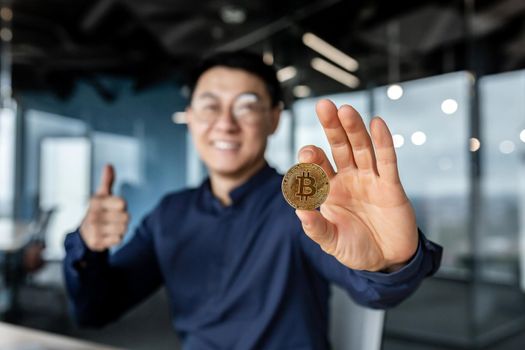 Close up photo of businessman's hand holding cryptocurrency money bitcoin, selective focus man smiling and happy showing thumbs up, working inside modern office building, successful investor.