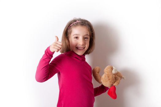 Portrait of a beautiful and smiling girl showing thumbs up while holding a teddy bear isolated on white