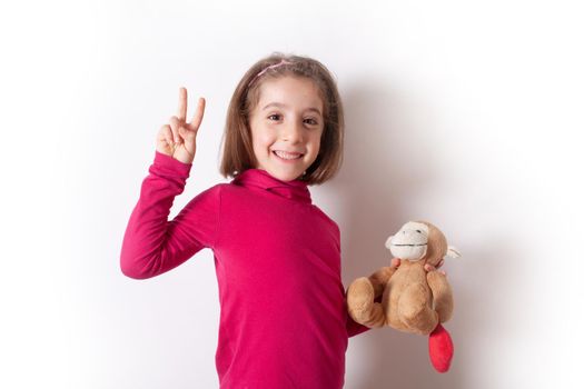 A cheerful little girl shows a victory sign while holding a Monkey Plush. The child is in a red pullover on a white background. happiness concept