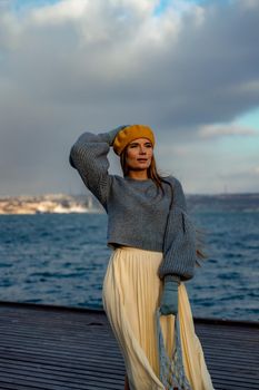 Outdoors fashion portrait of a beautiful middle aged woman walking on the beach. Marine background. Dressed in a stylish warm blue sweater, yellow skirt and beret