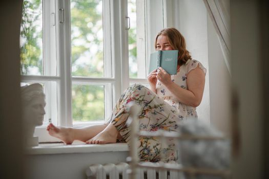 A middle-aged woman in a cream dress sits mysteriously and looks out the window on the windowsill. Green trees outside