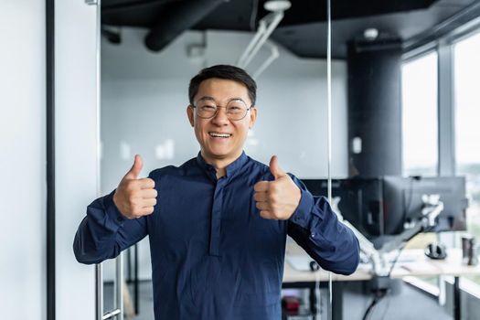 Portrait of successful asian businessman, man working in modern office looking at camera and smiling, broker trader showing thumbs up.
