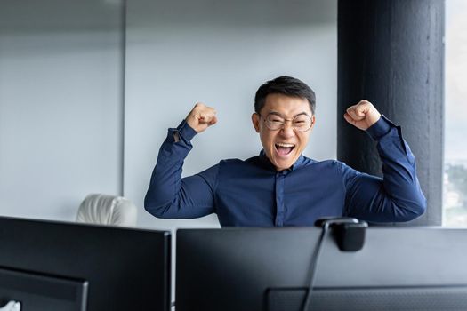 Happy Asian boss leader of programmer team, looking at computer screen and happy, celebrating victory, completed project, and submitted completed program, businessman entrepreneur holding hands up.
