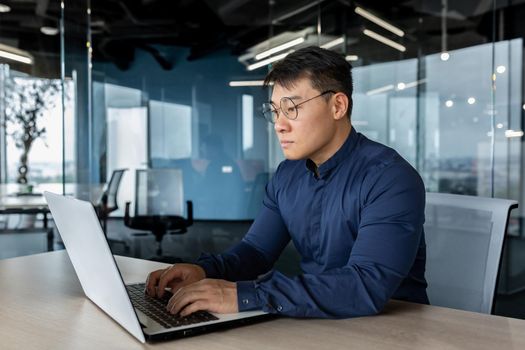 Serious and focused businessman working with laptop, man in glasses and shirt inside office building, asian boss typing on computer.