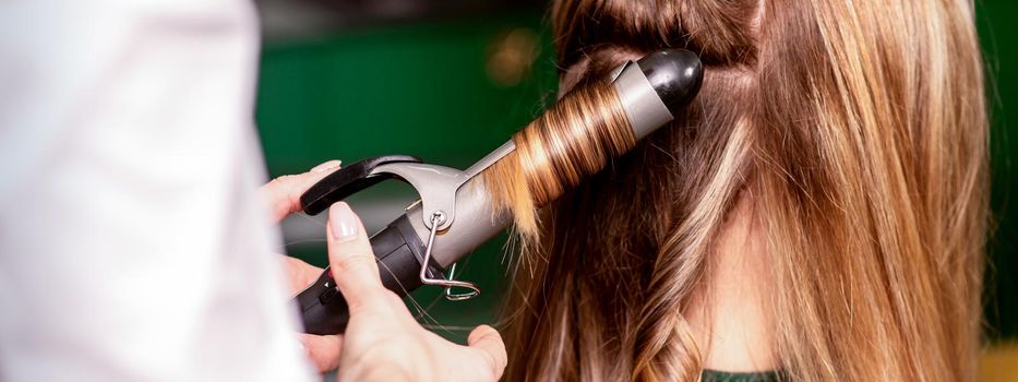 The female hairdresser is curling hair for a brown-haired young caucasian woman in a beauty salon