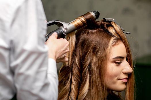 The female hairdresser is curling hair for a brown-haired young caucasian woman in a beauty salon