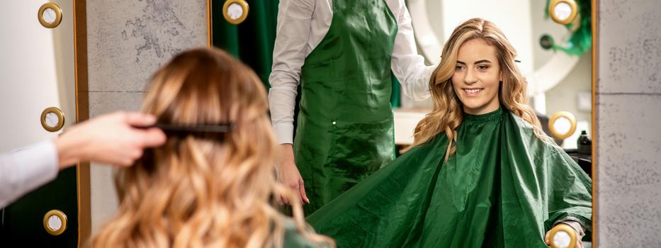 Hairdresser combing long hair of young caucasian woman looking and smiling in the mirror in beauty salon