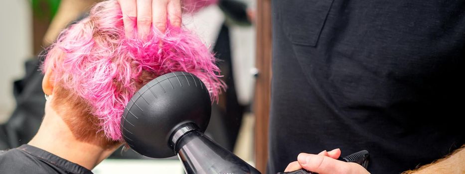 Drying short pink bob hairstyle of a young caucasian woman with a black hair dryer with the brush by hands of a male hairdresser in a hair salon, close up